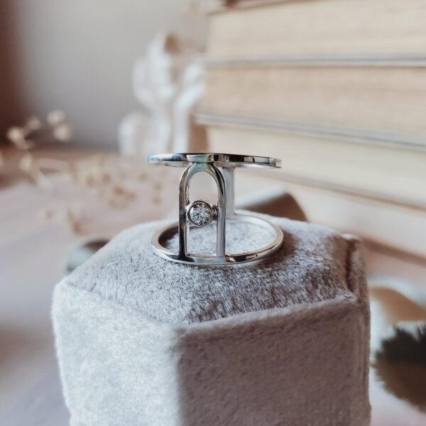 Faustina Wide Arch Statement Ring in sterling silver with central moissanite, resting on a cream velvet ring box surrounded by books and dried florals.