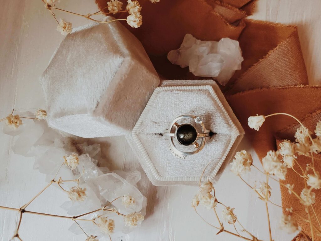 Luna y Terra black obsidian ring with a moissanite-accented encircling ring, displayed in a velvet ring box with ribbons and baby’s breath flowers.