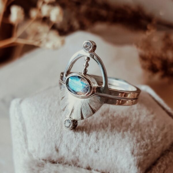 Pluto ring set with oval labradorite centerpiece, sterling silver interlocking design, displayed on cream velvet ring box with dried florals.