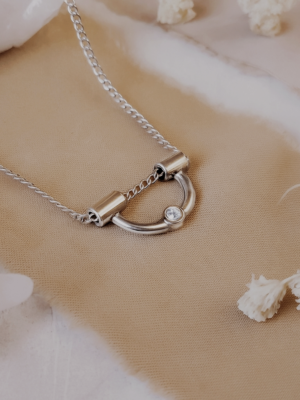 Tiny arch necklace in sterling silver with moissanite accent, displayed on neutral fabric with crystals and dried florals