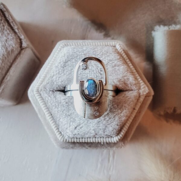Interlocking arch ring set in sterling silver with 14k yellow gold accent, featuring oval labradorite and moissanite accent stone, displayed in velvet ring box with sage green ribbon.