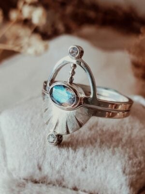 Pluto ring set with oval labradorite centerpiece, sterling silver interlocking design, displayed on cream velvet ring box with dried florals.