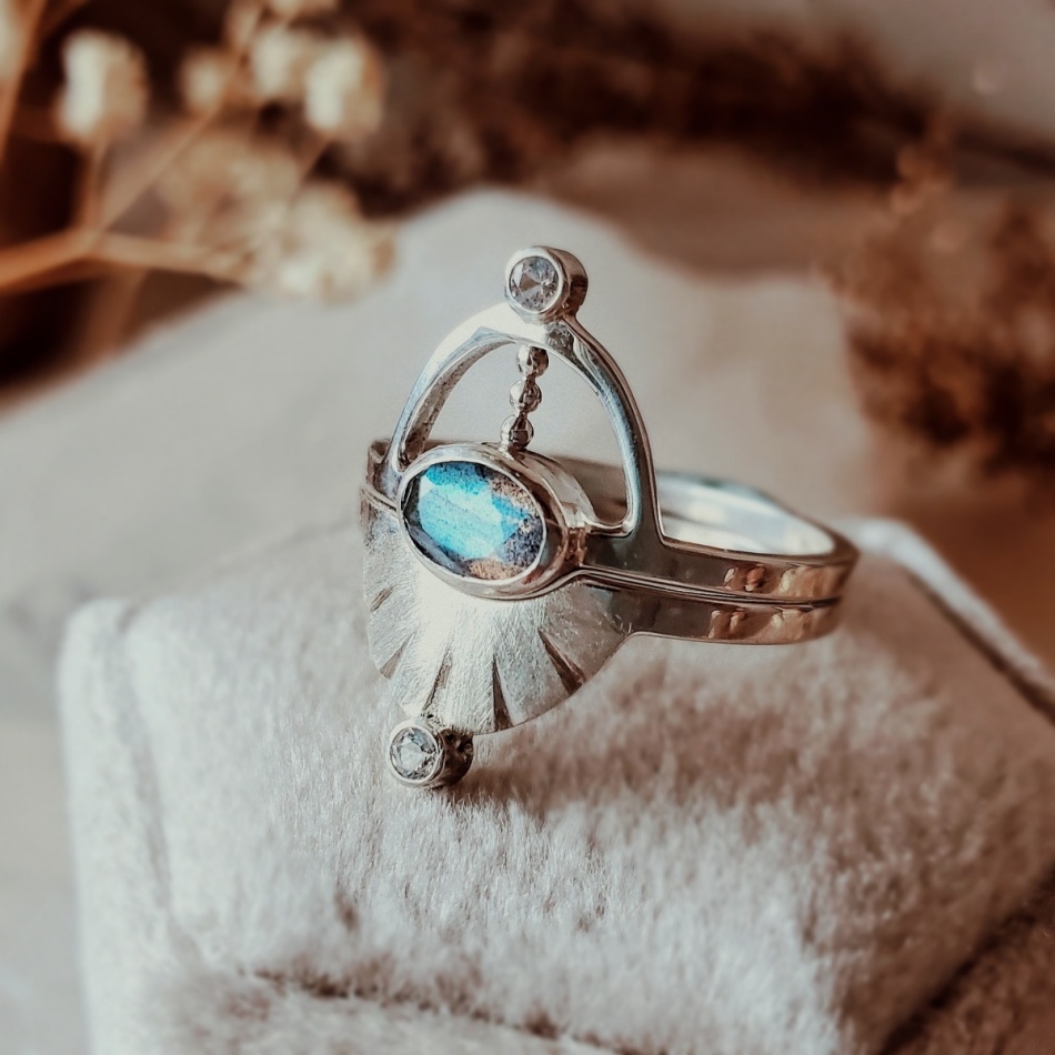 Pluto ring set with oval labradorite centerpiece, sterling silver interlocking design, displayed on cream velvet ring box with dried florals.
